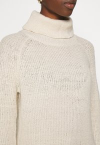 Cream-colored knit sweater with a high, folded collar, featuring a loose fit and visible textured weave.