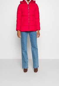 Doudoune rouge avec fermeture éclair à l'avant, capuche et deux poches latérales. Le tissu est lisse avec un motif matelassé. Associée à un jean bleu.