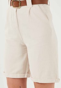 Pantaloni corti in cotone beige con vestibilità rilassata, con orlo arrotolato, tasche laterali e una cintura marrone con fibbia dorata. Texture liscia.