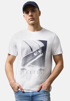 Junger Mann trägt ein weißes T-Shirt mit einem stilisierten Vintage-Autolicht-Design, blaue Jeans und eine schwarze Kappe.