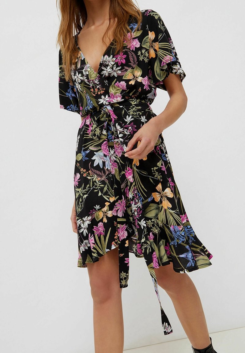 Femme portant une robe portefeuille noire à imprimé floral, manches courtes et taille nouée, se tenant devant un fond clair uni.