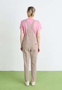 Pañales a rayas en color beige con un ajuste holgado, con tirantes y bolsillos. Combinados con una camiseta rosa y zapatillas blancas.