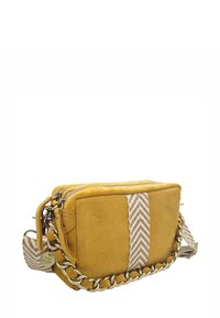 Sac bandoulière en daim jaune moutarde avec une bandoulière à motif chevrons, des accessoires en chaîne dorée et une fermeture zippée. Forme rectangulaire compacte.
