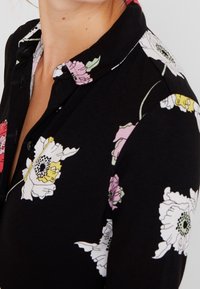 Black floral shirt with a collared neckline. Features large white and pink flowers with green accents. Soft fabric texture, button closure.