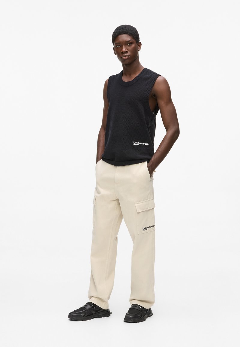 Débardeur noir sans manches en maille, pantalon cargo beige avec poches, baskets noires, modèle debout avec assurance devant un fond blanc.