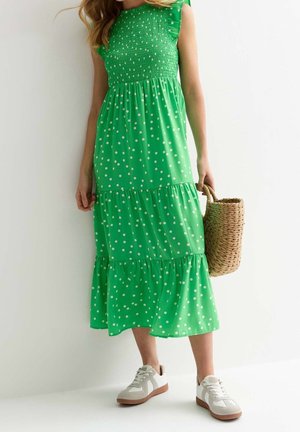 Žena v zelených bezrukávových midi šatech s puntíky a volánky, bílými teniskami a s pletenou slaměnou kabelkou.