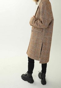 Ruskea ruudullinen takki, jossa on suora leikkaus, sivuleikkaukset ja pehmeä rakenne, yhdistettynä mustiin legginsseihin ja paksupohjaisiin mustiin saappaisiin.