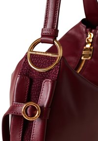 Borsa a tracolla in pelle bordeaux con hardware dorato, accento in tessuto testurizzato e un rinforzo circolare dorato ben visibile sulla tracolla.