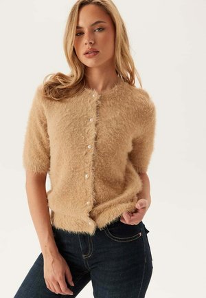 Kvinna som bär en beige fluffig kortärmad knäpptröja och mörkblå jeans, poserar med en hand i fickan.