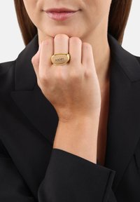 Anello color oro con forma ovale, caratterizzato da un'incisione di "¡OOOP!" su una superficie liscia, indossato su una mano su uno sfondo nero.