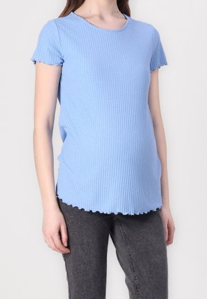 Kobieta w jasnoniebieskiej, prążkowanej bluzce z krótkim rękawem i ciemnoszarych dżinsach, stojąca na tle gładkiej, białej ściany.