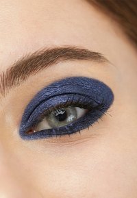Mörkblå texturerad ögonskugga applicerad på ögonlocket, med definierad eyeliner och långa ögonfransar som framhäver en ljusfärgad iris.