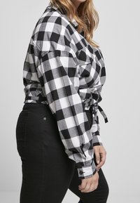 Donna in camicia a maniche lunghe a quadri bianchi e neri legata in vita, che indossa pantaloni neri aderenti, in piedi di lato contro uno sfondo neutro.