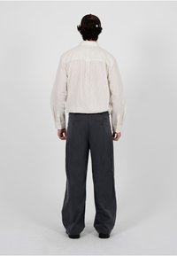 Homme portant une casquette marron foncé, une chemise rayée crème à manches longues et un pantalon ample gris foncé, debout de dos sur un fond uni.