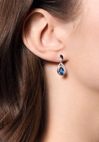 Srebrne kolczyki w kształcie kropli z niebieskim kamieniem w kształcie łzy, otoczone małymi przezroczystymi kamieniami, z zakręconym wzorem i polerowanym wykończeniem.
