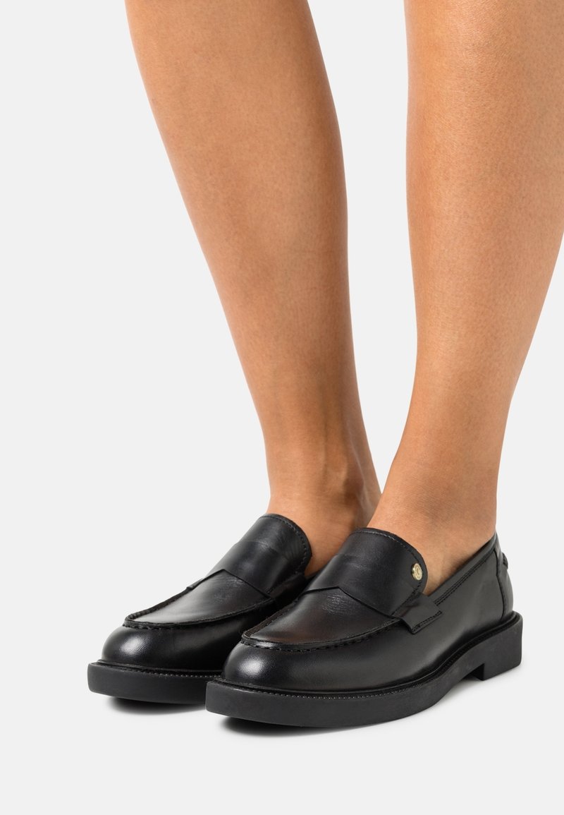 Mocassins en cuir noir portés pieds nus avec des semelles basses et épaisses et un bouton doré décoratif sur le côté.