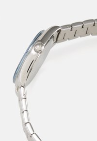 Orologio in acciaio inossidabile argentato con lunetta blu rotonda, corona testurizzata e bracciale a maglie, che mostra un design elegante e lucido.