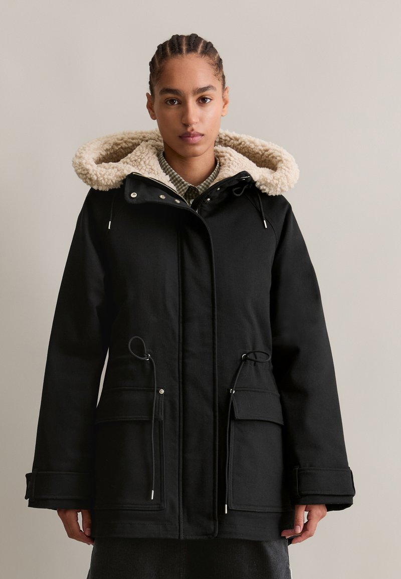 Zwarte parka met een beige fleece-voering in de capuchon. Voorzien van steekzakken aan de voorkant, een taille met trekkoord en een ritsafsluiting. Gemaakt van een gestructureerde stof.