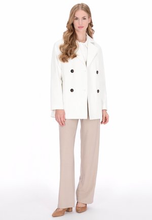 Witte dubbelgeknoopte jas met zwarte knopen, gecombineerd met beige broek en beige hakken, gepresenteerd op een model met golvend haar.