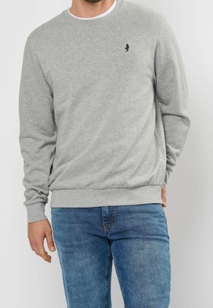 Mann trägt hellgrauen langärmeligen Sweatshirt mit kleinem dunklem Logo auf der Brust und blaue Jeans und steht vor einfarbigem Hintergrund.