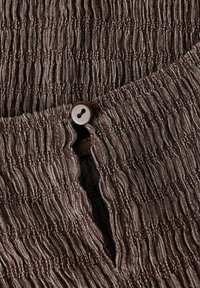 Tissu brun texturé avec des plis verticaux, orné d'un bouton marron à l'ouverture. Le matériau présente un léger éclat et une légère élasticité.