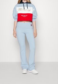 Kort sweatshirt i ljusblått med röd nedre del och guldlogo; kombinerad med ljusblå utsvängda byxor och vita skor.