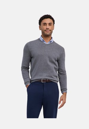 Mann trägt einen grauen V-Ausschnitt-Pullover über einem hellblauen Hemd mit Kragen, marineblaue Hose und braunen Gürtel, steht mit einer Hand in der Tasche.