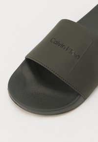 Zeleni gumijasti natikač z teksturiranim podplatom in gladkim trakom, ki ima viden "Calvin Klein" logotip na zgornji površini.