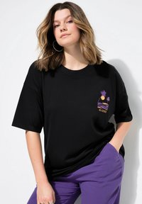 Černé oversized tričko s grafikou, která zobrazuje slunce, vlny a palmy v purpurové a oranžové barvě, doplněné o texturované purpurové kalhoty.