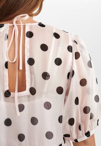 Genomskinlig, lätt blus i mjuk rosa med stora svarta prickar. Har en nyckelhål baktill med bandknut och puffärmar.