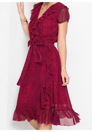 Donna che indossa un vestito con stampa leopardata rossa e nera con volant, maniche corte e cintura annodata in vita, posando su sfondo bianco.