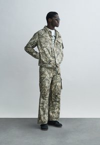 Σετ καμουφλαρισμένο με σακάκι και παντελόνι σε τόνους καφέ και πράσινου, κατασκευασμένο από υφασμάτινο με υφή. Μαύρα υποδήματα. Λευκό T-shirt με γραφικό σχέδιο από κάτω.