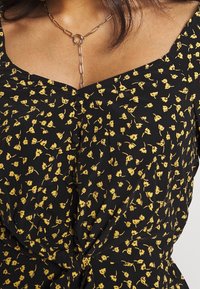 Czarna sukienka z kwiatowym nadrukiem w żółte kwiaty, z dekoltem w kształcie serca i węzłem w pasie. Akcent w postaci naszyjnika z łańcuchem w kolorze złotym.