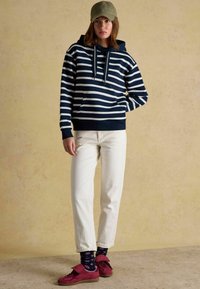 Žena v zelenej čiapke, modro-bielo pruhovanom mikine, bielych nohaviciach, vzorovaných tmavých ponožkách a červených teniskách stojí pred béžovým pozadím.