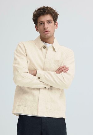 Junger Mann mit lockigem Haar, der eine beigefarbene, geknöpfte Jacke und dunkle Hosen trägt und mit verschränkten Armen vor einem einfarbigen Hintergrund steht.