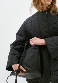 Giacca nera trapuntata con colletto alto, caratterizzata da motivi triangolari. La persona tiene una borsa a tracolla in pelle nera con un accento annodato.