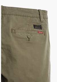 Poche arrière d'un pantalon Khaki Levi's avec fermeture à bouton et deux étiquettes en tissu de la marque au-dessus de la poche sur fond blanc.