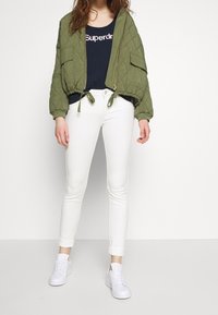 Chaqueta bomber acolchada verde oliva con cremallera y detalle de lazo, combinada con jeans ajustados blancos y zapatillas blancas, con una camiseta de tirantes azul marino.