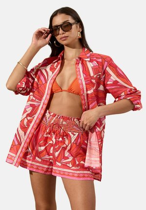 Naine kannab üle suuruse punase ja roosa mustriga särki ning sobivaid lühikesi pükse, oranži bikiinitopiga ja suuri päikeseprille ning reguleerib prille.