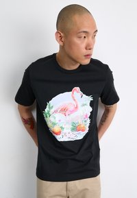 T-shirt nera con una grafica colorata di un fenicottero circondato da piante tropicali e frutta, con un elemento di design rotondo.
