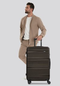 Man in een beige outfit die de handgreep vasthoudt van een grote zwarte hard-shell koffer met een gestructureerd horizontaal ribbelpatroon op wielen.