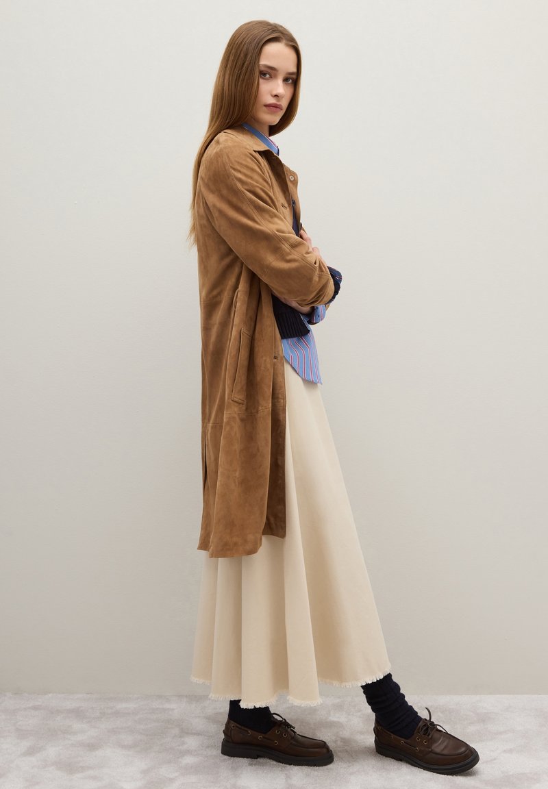 Cappotto lungo in suede marrone sopra una camicia a righe blu e bianche, abbinato a una gonna color crema e scarpe marroni, con calzini blu navy a coste.