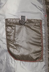 Mörkgrå tygficka med en svart knapp, säkrad av en randig flik. Slät yta med röda sömnadsdetaljer längs kanterna.