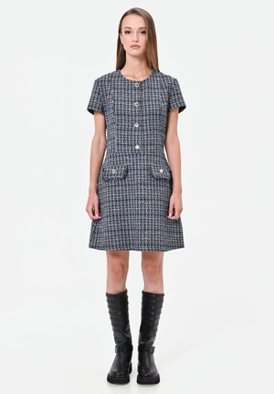 Donna che indossa un vestito in tweed a maniche corte, abbottonato, con tasche frontali e stivali neri al ginocchio, in piedi davanti a uno sfondo neutro.