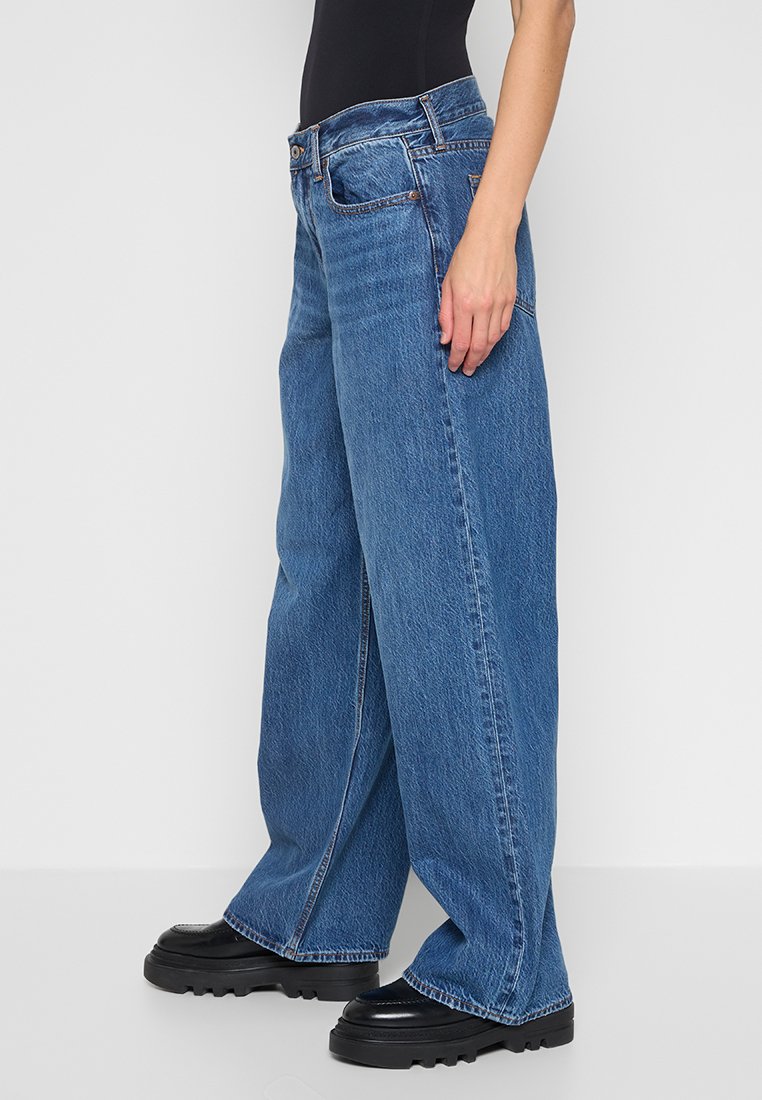 Weite blaue Denim-Jeans mit hoher Taille, die ein traditionelles Fünf-Taschen-Design und dezente Ausbleichungen aufweisen. Begleitet von schwarzen klobigen Schuhen.