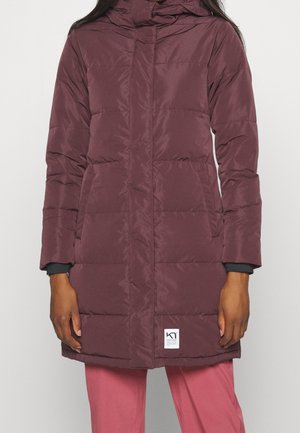 Kobieta ubrana w długi, pikowany, bordowy płaszcz zimowy z kapturem i bocznymi kieszeniami, w połączeniu z różowymi spodniami.