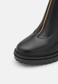 Bottine noire en cuir avec fermeture éclair avant dorée et semelle épaisse à crampons, vue de face en gros plan sur fond blanc.