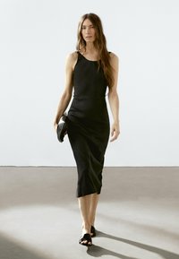 Vestito midi nero senza maniche con silouette aderente e spacco laterale, abbinato a sandali neri aperti sulla punta e una piccola borsa nera.
