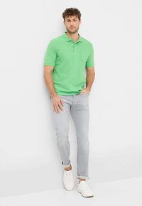 Polo verde a maniche corte con taschino, abbinato a jeans grigio chiaro e sneakers bianche, su uno sfondo semplice.