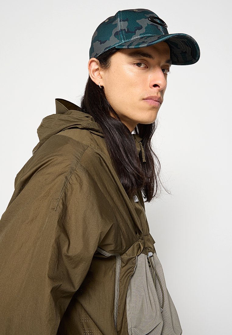 Chaqueta ligera marrón con capucha, con un bolsillo de malla y detalles de botones. Gorro de camuflaje azul con visera curvada.